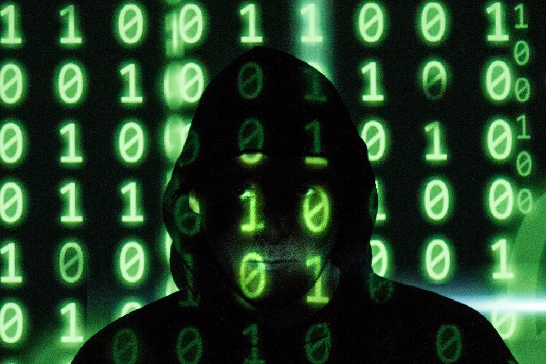 Hacker férfi áll a sötétben, az arcára bináris kód vetítve zöld színnel.